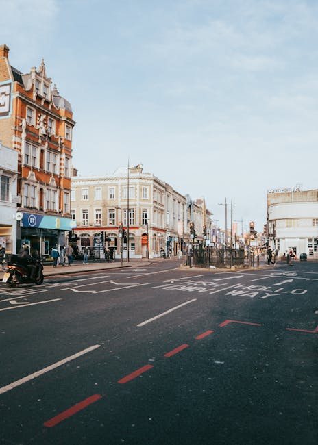 The image depicts a busy urban street scene with a mixture of historic and modern architecture lining the street, including brick and cream-colored buildings with ornate facades and large windows. The